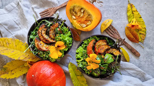 Zwei Schalen mit grünem Herbstsalat, ofengerösteten Hokkaido-Kürbisstücken und essbaren Blüten, serviert in handgefertigten Kokosnuss-Schalen auf einem herbstlich dekorierten Tisch mit Kürbis und bunten Blättern.