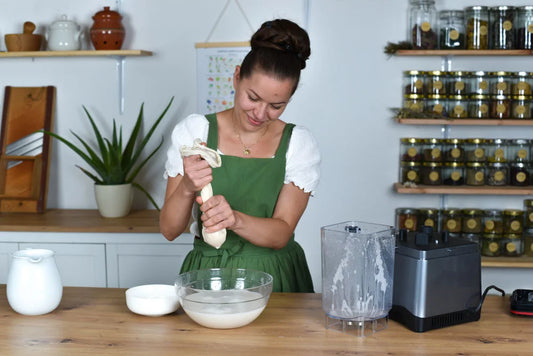 Frau presst mit einem Baumwoll-Nussmilchbeutel frische Hafermilch in eine Glasschüssel in ihrer Küche aus – nachhaltige, plastikfreie Zubereitung von pflanzlicher Milch.