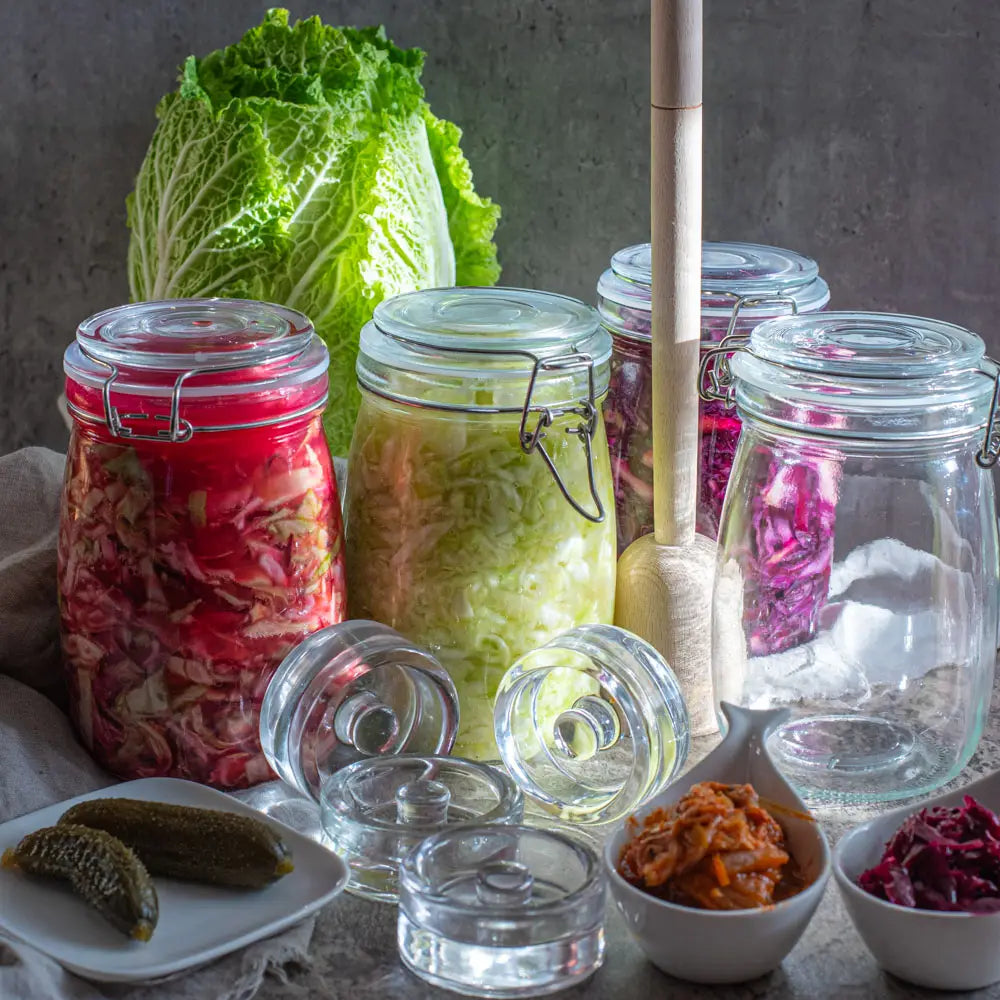 Fermentiertes Gemuese in 1L Buegelglaesern mit Glasgewichten und Holzstampfer auf einem Steintisch ideal fuer Sauerkraut und Kimchi zuhause.