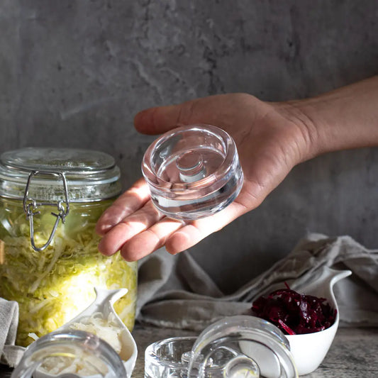 Hand hält ein Glasgewicht für Fermentation, daneben Sauerkraut im Glas und rote Bete im Schälchen.