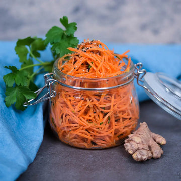 Fermentierter Karottensalat im Glas mit Ingwer, fein gehobelten Karotten und Gewürzen, angerichtet auf grauer Steinfläche mit blauem Leinentuch und frischen Kräutern.