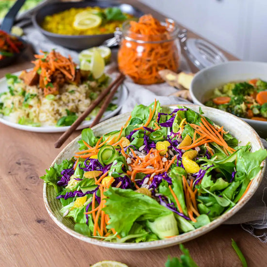 Bunter veganer Krautsalat mit Rotkraut, Karotten, Blattsalat und Ingwer, angerichtet in Keramikschale auf Holztisch, umgeben von Bowls, Suppe und fermentierten Karotten.