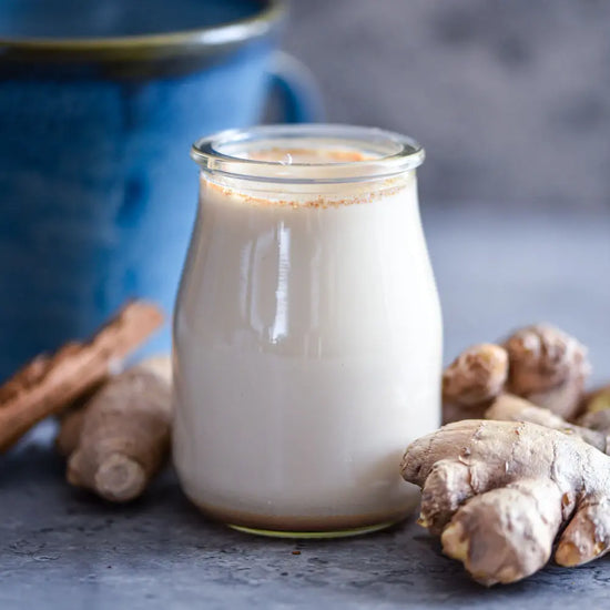 Vegane goldene Ingwer-Milch im Glas, umgeben von frischer Ingwerwurzel und Gewürzen, auf grauer Steinfläche mit blauer Keramik Tasse im Hintergrund.
