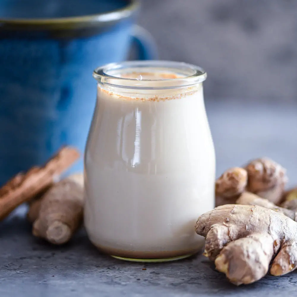 Vegane goldene Ingwer-Milch im Glas, umgeben von frischer Ingwerwurzel und Gewürzen, auf grauer Steinfläche mit blauer Keramik Tasse im Hintergrund.