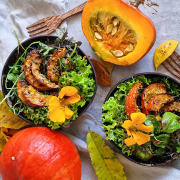 Zwei Schalen mit grünem Herbstsalat, ofengerösteten Hokkaido-Kürbisstücken und essbaren Blüten, serviert in handgefertigten Kokosnuss-Schalen auf einem herbstlich dekorierten Tisch mit Kürbis und bunten Blättern.