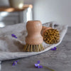 Holzbürste mit Naturborsten auf Stoff und grauer Oberfläche mit kleinen violetten Blüten – ideal zum Reinigen von Geschirr und Pfannen.