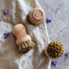 Mehrere Holzbürsten mit Naturborsten auf Stoff und grauer Oberfläche mit kleinen violetten Blüten – ideal für Küche und Haushalt.