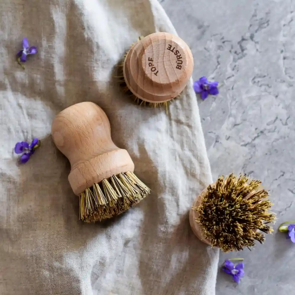 Mehrere Holzbürsten mit Naturborsten auf Stoff und grauer Oberfläche mit kleinen violetten Blüten – ideal für Küche und Haushalt.