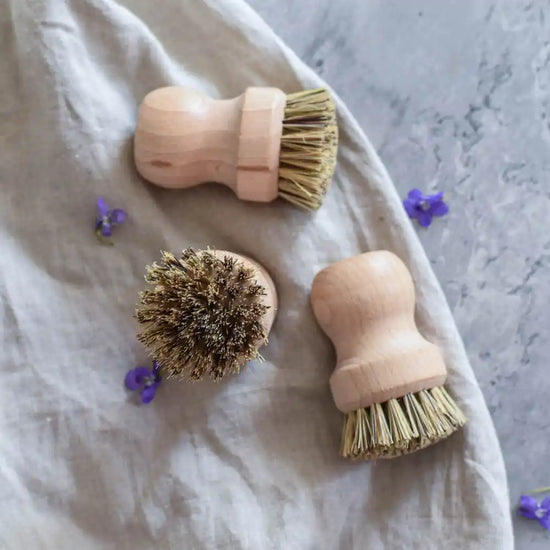 Mehrere Holzbürsten mit Naturborsten auf Stoff und grauer Oberfläche mit kleinen violetten Blüten – ideal für Küche und Haushalt.