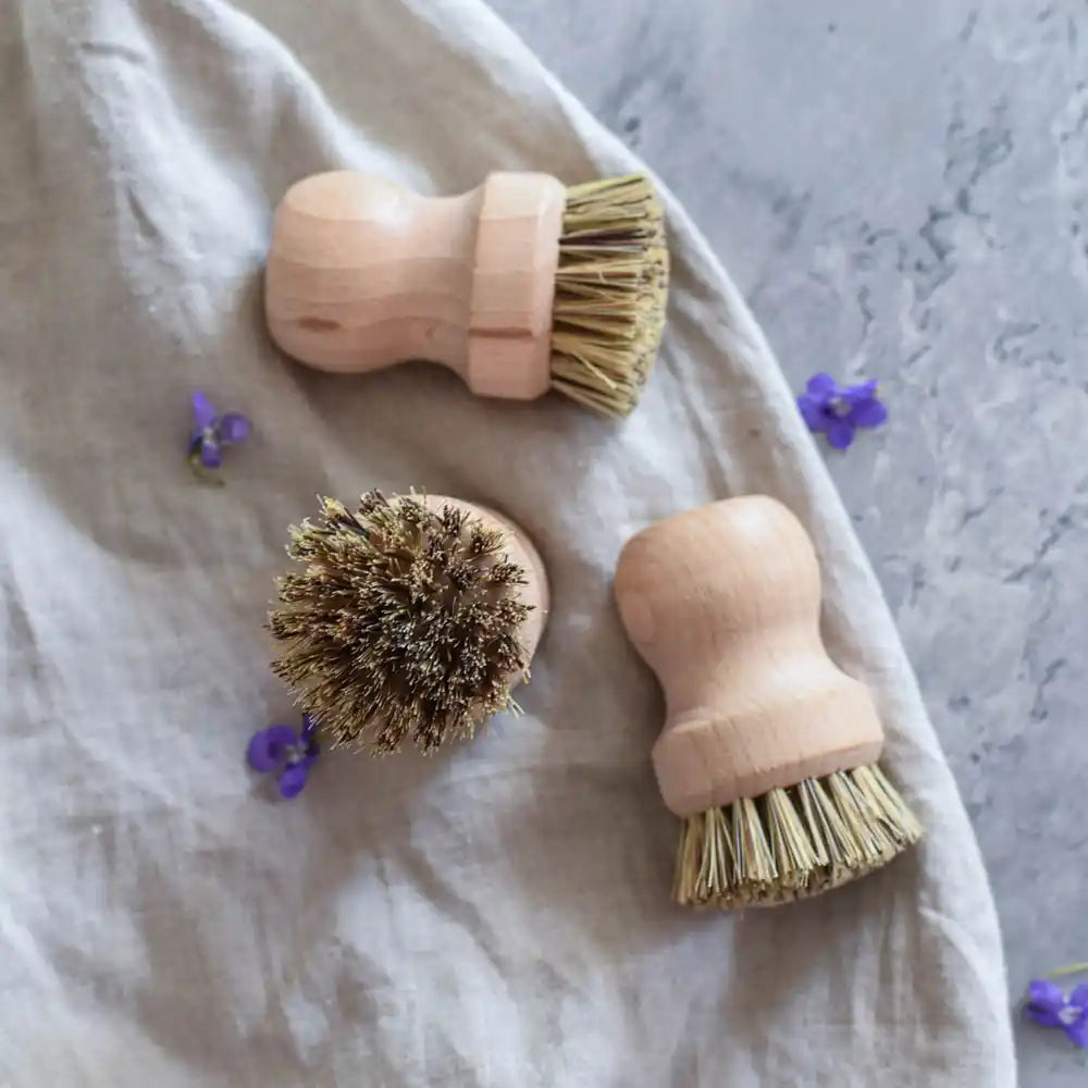 Mehrere Holzbürsten mit Naturborsten auf Stoff und grauer Oberfläche mit kleinen violetten Blüten – ideal für Küche und Haushalt.