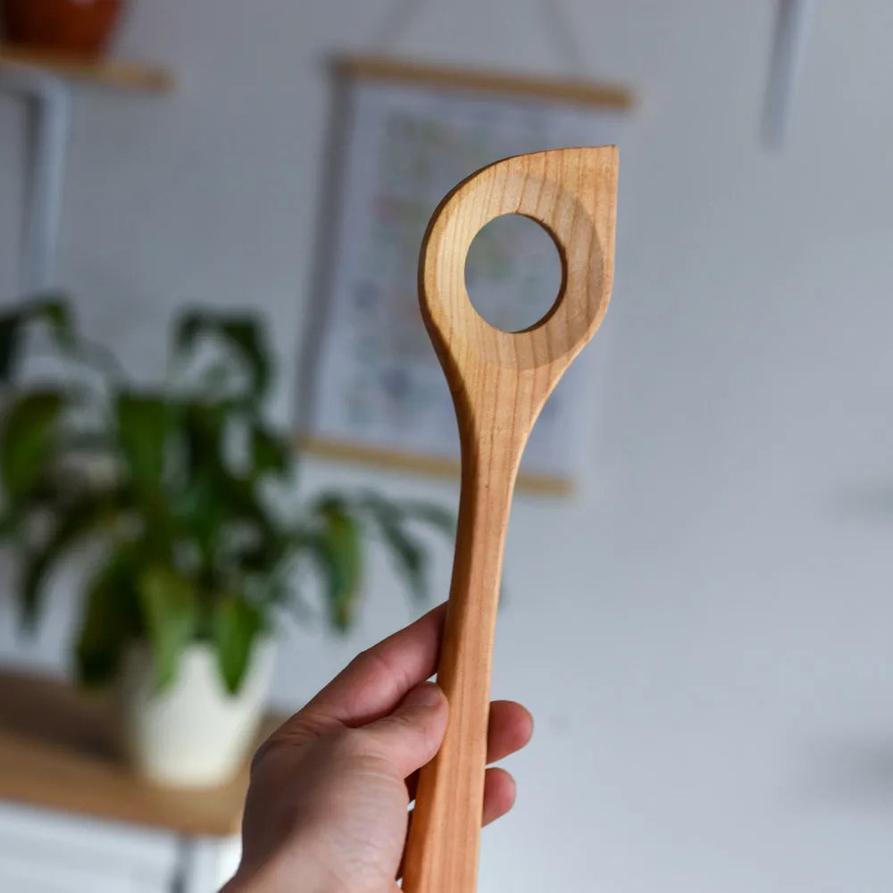 Holzkochlöffel mit Loch wird in der Hand gehalten vor unscharfem Hintergrund mit Pflanze – ideal zum Kochen und Umrühren in der Küche.