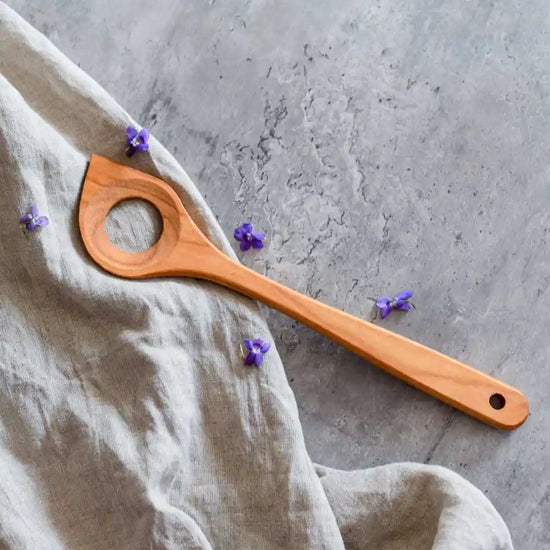 Holzkochlöffel mit Loch auf Stoff und grauer Oberfläche mit kleinen violetten Blüten – ideal zum Kochen und Umrühren in der Küche.