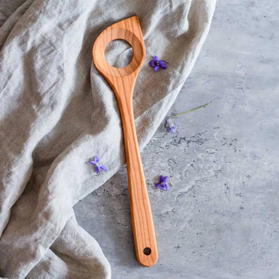 Holzkochlöffel mit Loch auf Stoff und grauer Oberfläche mit kleinen violetten Blüten – ideal zum Umrühren und Kochen in der Küche.