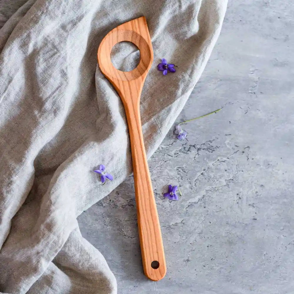 Holzkochlöffel mit Loch auf Stoff und grauer Oberfläche mit kleinen violetten Blüten – ideal zum Umrühren und Kochen in der Küche.