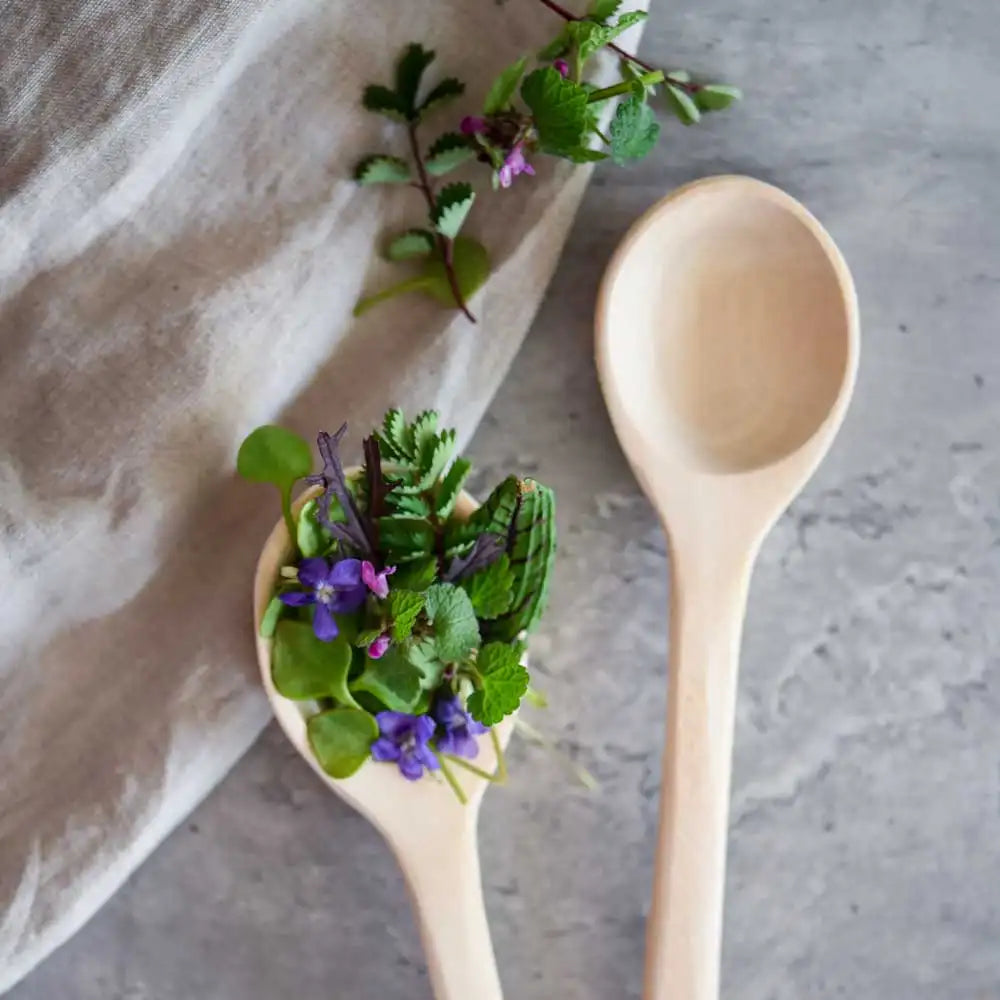 Zwei Holzlöffel, einer gefüllt mit Wildkräutern und Blüten auf Stoff – ideal zum Servieren und Anrichten.