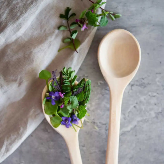 Zwei Holzlöffel, einer gefüllt mit Wildkräutern und Blüten auf Stoff – ideal zum Servieren und Anrichten.