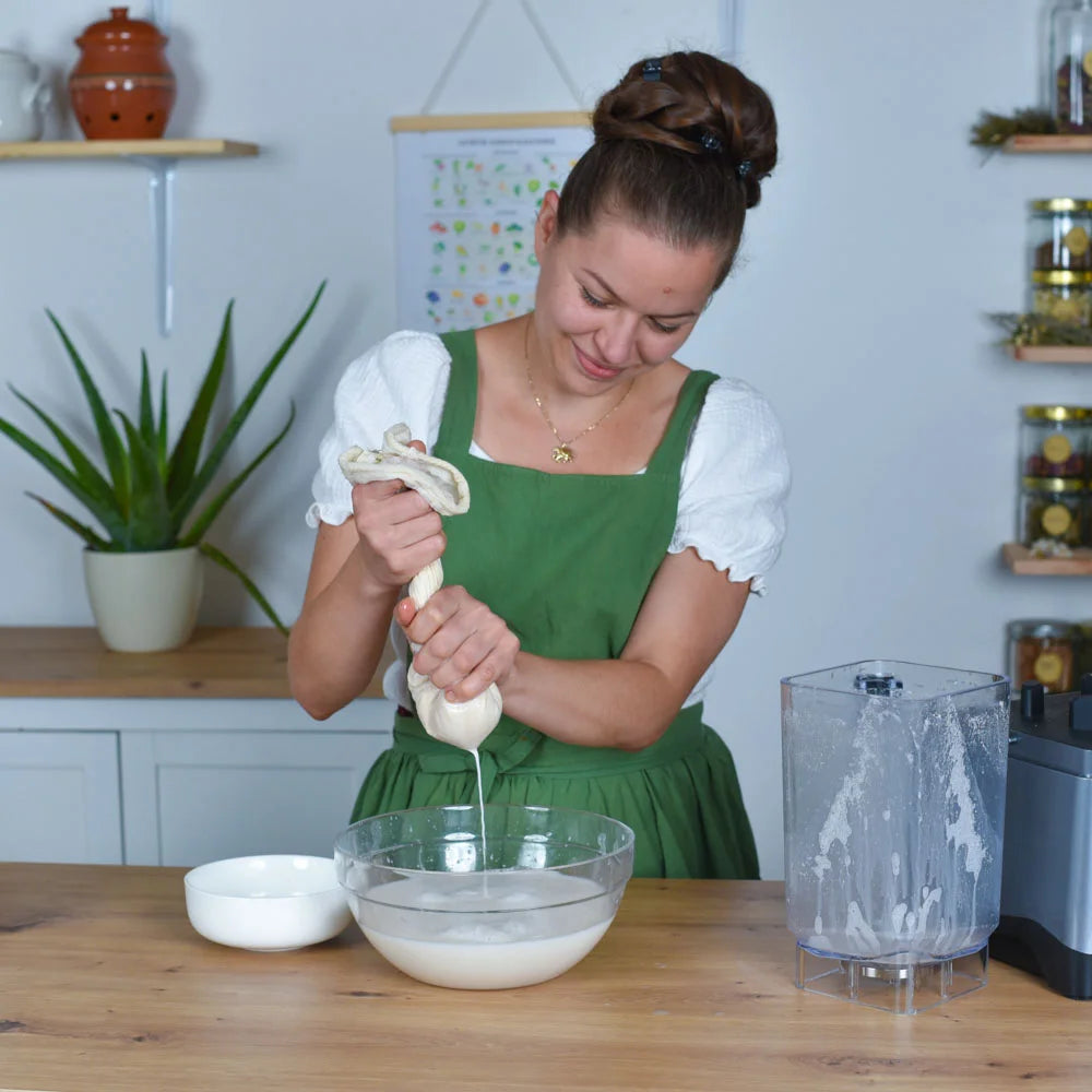 Eine Frau presst Mandelmilch durch einen Baumwoll-Nussmilchbeutel über einer Glasschüssel in einer Küche.