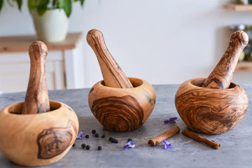 Drei Mörser mit Stößeln aus Olivenholz auf grauer Oberfläche mit Pfefferkörnern, Zimtstangen und Blüten – stilvoll und praktisch in der Küche.