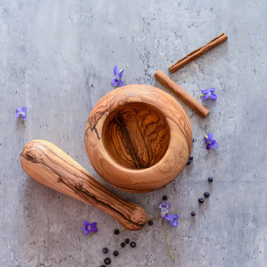 Olivenholz Mörser mit Stößel, Pfefferkörnern, Zimtstangen und kleinen violetten Blüten auf grauer Oberfläche – natürliches Küchenutensil.