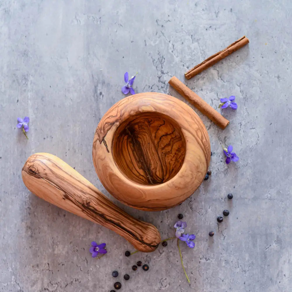 Olivenholz Mörser mit Stößel, Pfefferkörnern, Zimtstangen und kleinen violetten Blüten auf grauer Oberfläche – natürliches Küchenutensil.