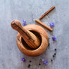 Mörser und Stößel aus Olivenholz mit Pfefferkörnern, Zimtstangen und violetten Blüten auf grauer Oberfläche – dekorativ und praktisch.