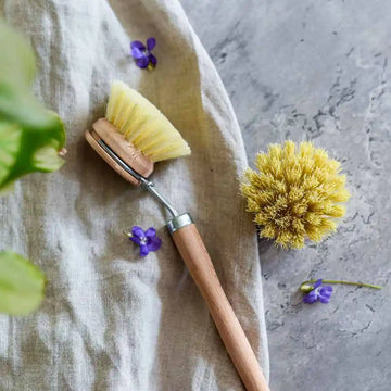 Spülbürste aus Holz mit Wechselkopf und Naturborsten auf Stoff mit kleinen violetten Blüten – ideal für Küche und Haushalt.