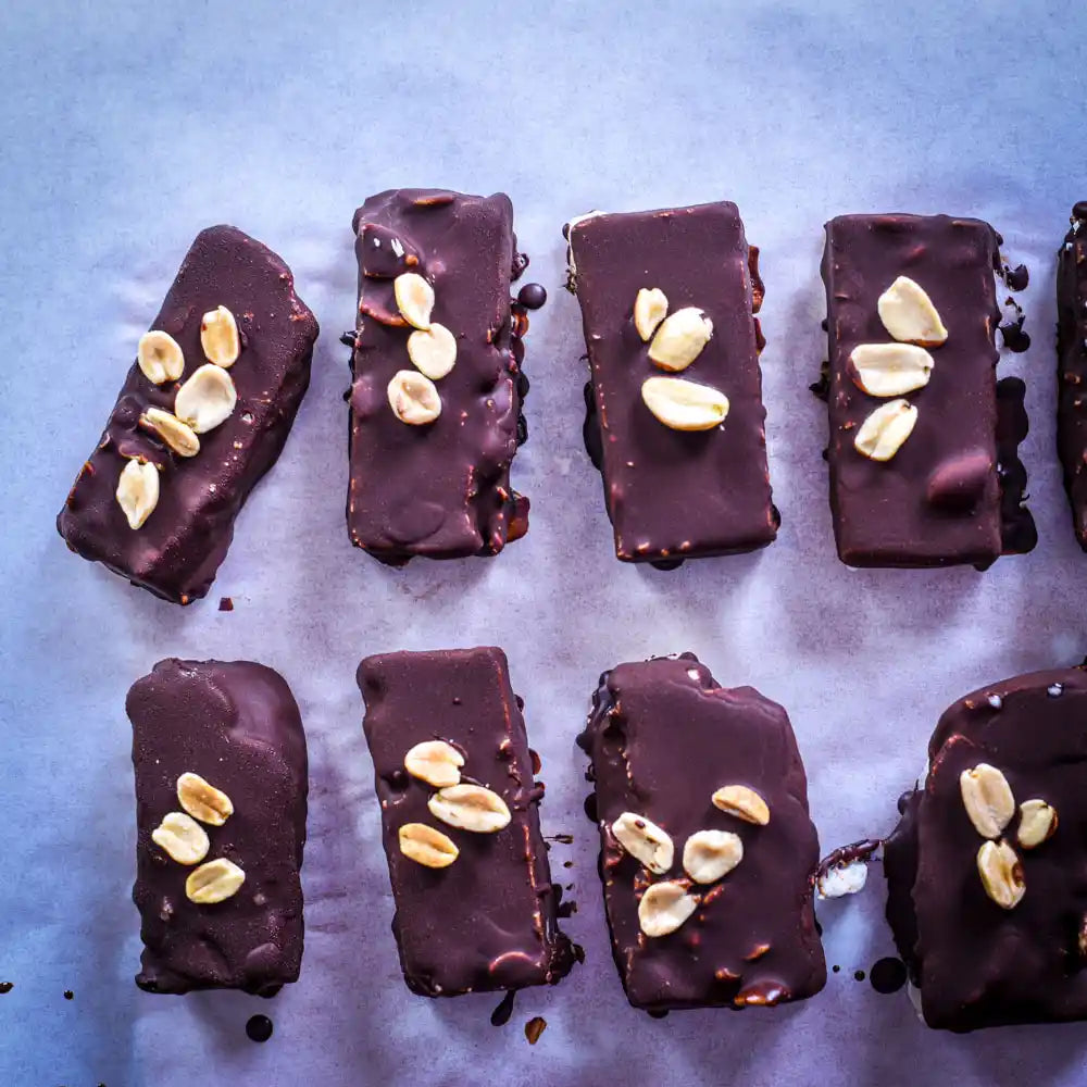 Vegane Eisriegel mit dunkler Schokolade, Erdnüssen und cremiger Erdnuss-Karamell-Füllung auf Backpapier.