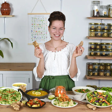 Frau in heller Naturküche hält frischen Ingwer in den Händen, vor ihr vegane Gerichte wie Suppe, Bowls und Salate, im Hintergrund Regale mit Fermentgläsern und ruhige schöne Küchenatmosphäre.