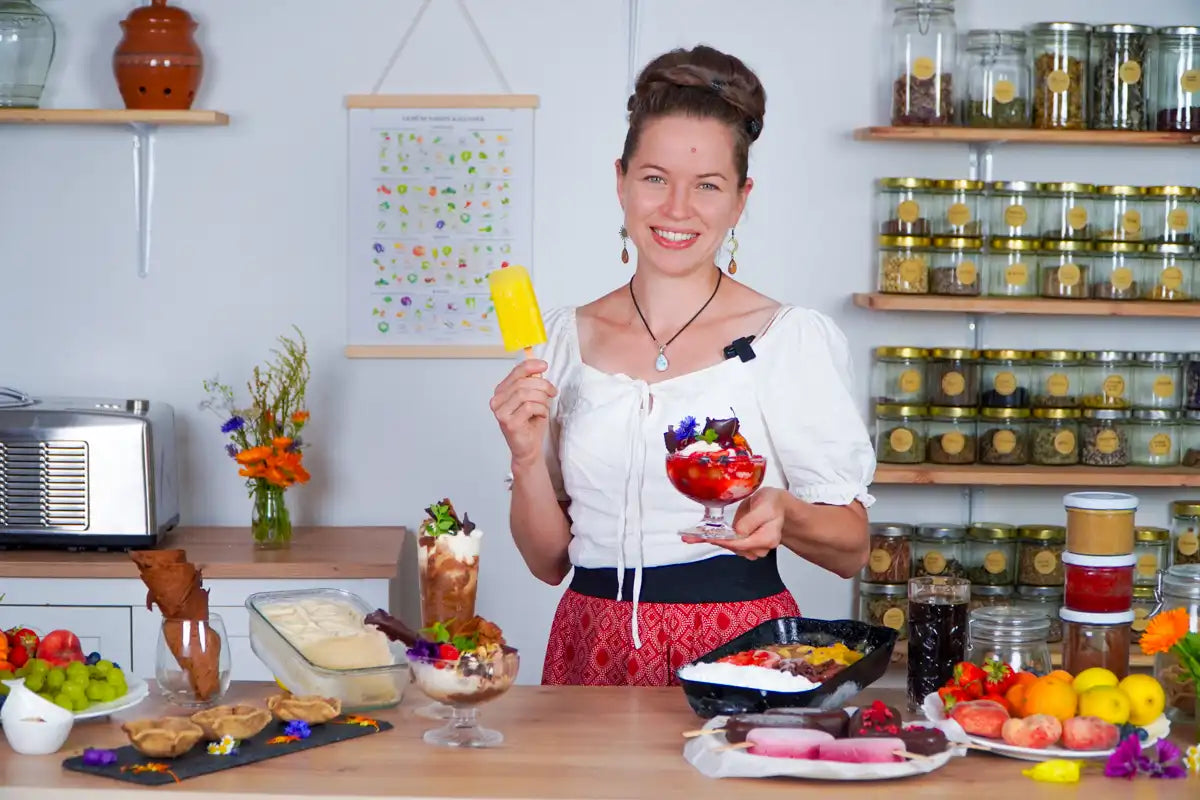 Lächelnde Frau präsentiert gelbes Eis am Stiel und einen bunten veganen Eisbecher vor vielen Eiskreationen.