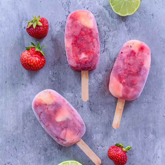 Drei vegane Erdbeer-Limetten-Eis am Stiel mit Erdbeerstückchen, frischen Erdbeeren und Limetten auf grauem Untergrund.