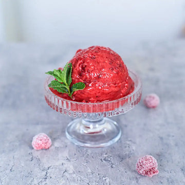 Rotes veganes Himbeersorbet mit Minze in einer Glasschale, umgeben von gefrorenen Himbeeren.