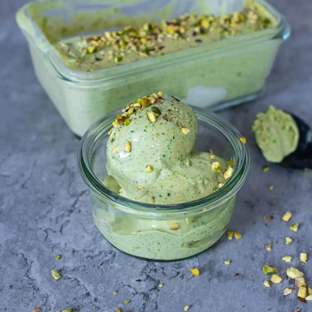 Cremiges veganes Pistazieneis im Glas, garniert mit gehackten Pistazien und einer Eisbox im Hintergrund.