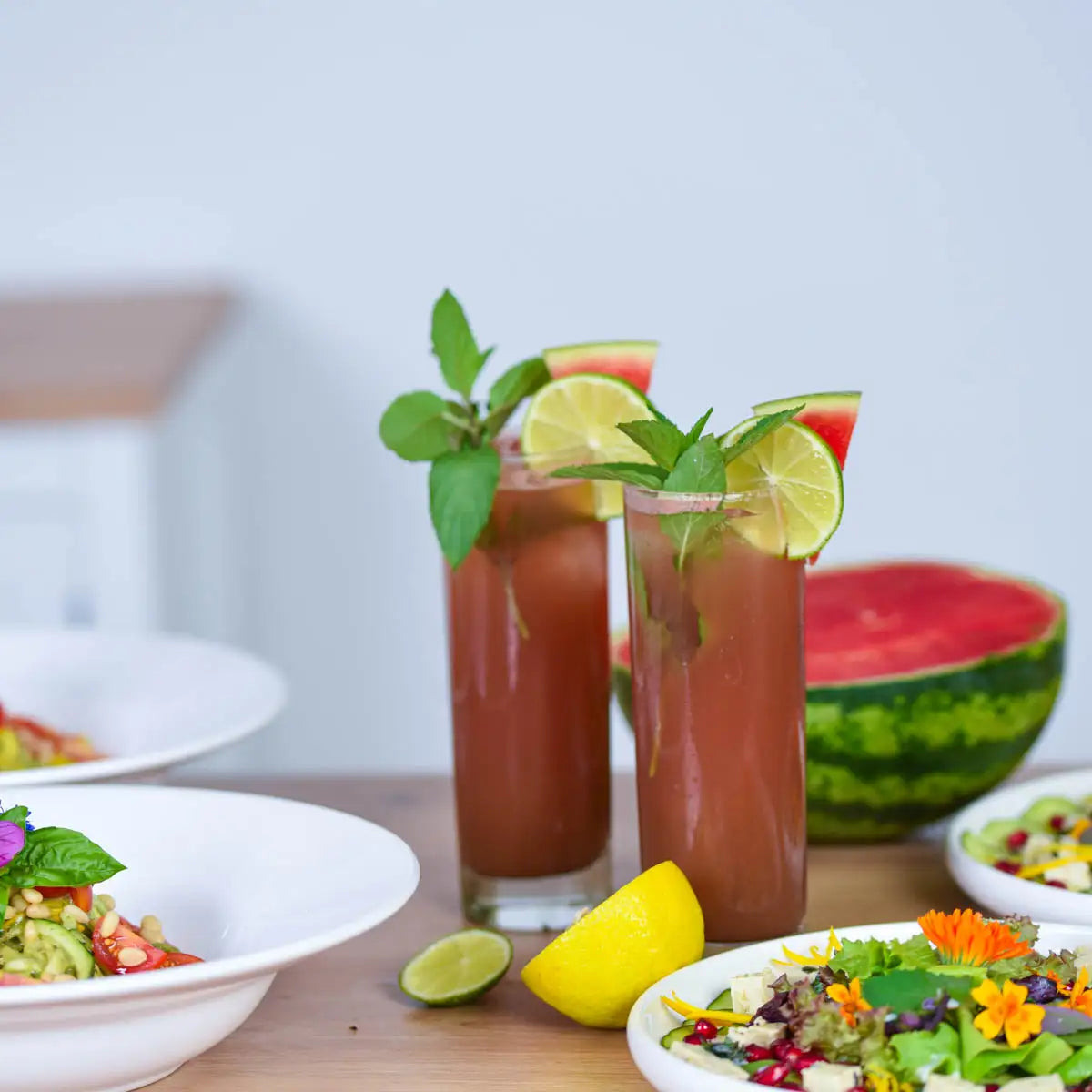 Zwei Gläser mit rotem Wassermelonen-Getränk, Limette und Minze dekoriert, im Hintergrund Salate und Wassermelone.