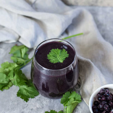 Dunkelvioletter Beeren-Smoothie in einem Glas, garniert mit frischen Kräutern. Daneben liegen gefrorene Beeren und grüne Wildkräuter auf hellem Hintergrund. Antioxidantienreicher Smoothie für eine pflanzliche, vitaminreiche und natürliche Ernährung.