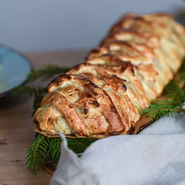 Goldbraune Blätterteigrolle mit sternförmigem Muster, auf einem Holzbrett und umgeben von Tannengrün, seitliche Detailansicht.