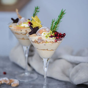 Zwei Dessertgläser mit cremigen Schichten, dekoriert mit Nüssen, Granatapfelkernen, Gebäck und einem kleinen Tannenzweig.