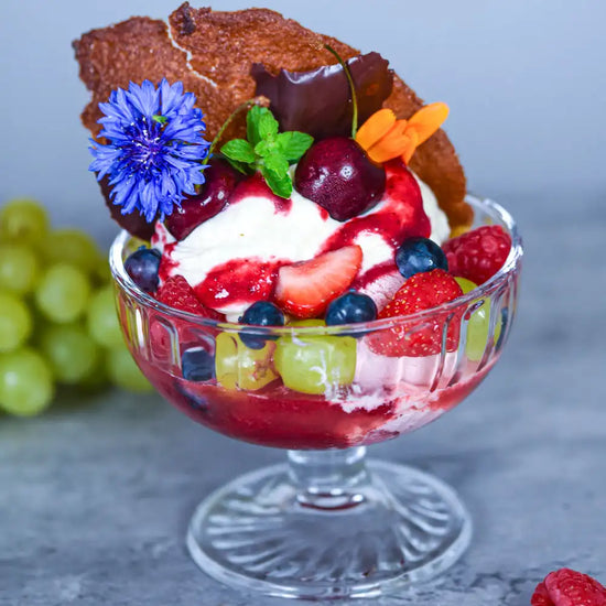 Glasschale mit Vanilleeis, Beeren, Kirschen, Trauben, Karamell-Chip und blauer Kornblume dekoriert.