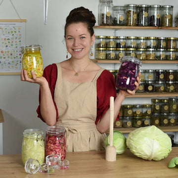 Theresa haelt zwei Fermentationsglaeser mit gelbem und violettem Gemuese, vor ihr stehen weitere Gläser, Glasgewichte und frischer Kohl.