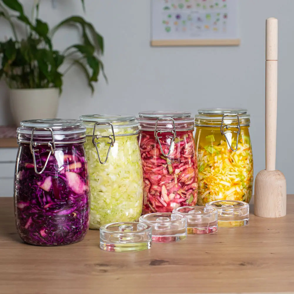 Vier 1-Liter-Fermentationsglaeser mit verschiedenem Gemuese stehen neben Glasgewichten und einem Holzstampfer auf dem Tisch.