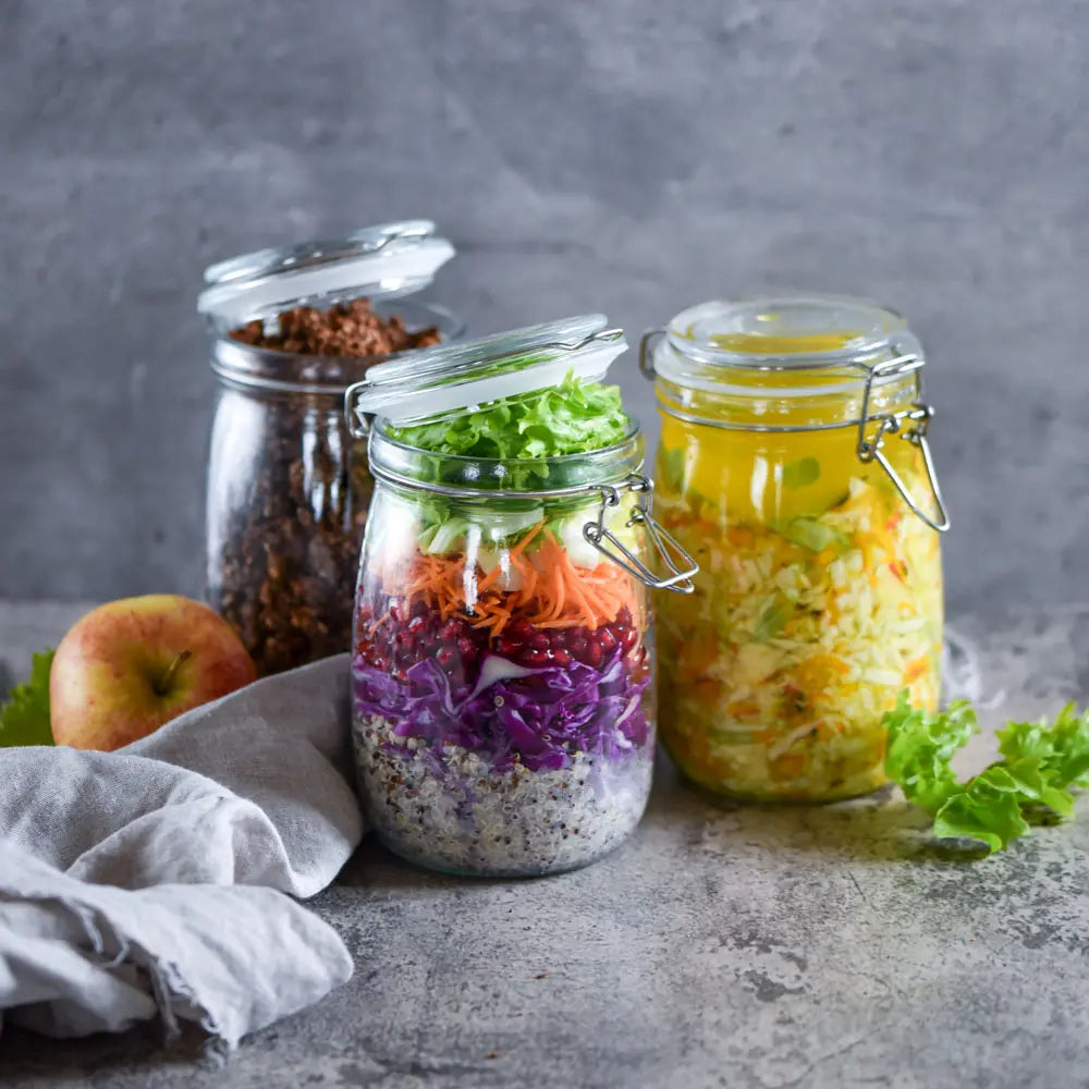 Drei Bügelgläser mit Schichtsalat, fermentiertem Krautsalat und Granola stehen auf einem grauen Untergrund neben Apfel und Tuch.