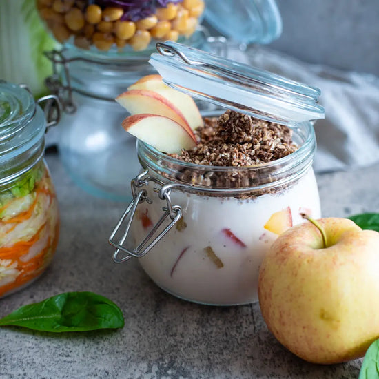 Ein 500-ml-Buegelglas ist mit veganen Overnight-Oats, Apfelstuecken und Granola gefuellt; daneben liegen frischer Apfel und Spinatblaetter.