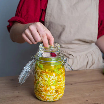 Eine Hand setzt ein Glasgewicht auf fermentiertes Gemüse im 1-Liter-Drahtbügelglas, um das Kraut sicher unter der Lake zu halten.