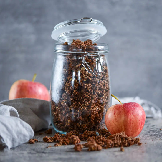 Offenes Glas mit schokoladigem Granola, daneben frische Äpfel und verstreute Müslostücke auf grauem Untergrund.