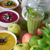 Grüner Wildkräuter-Smoothie mit Äpfeln und Limette neben bunten Gemüsesuppen für eine gesunde Ernährung.