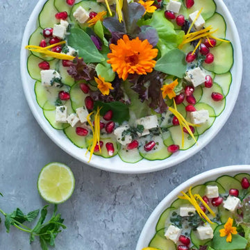 Draufsicht auf zwei Teller Gurken-Carpaccio mit Feta, Granatapfelkernen, Blattsalat und Blüten dekoriert.