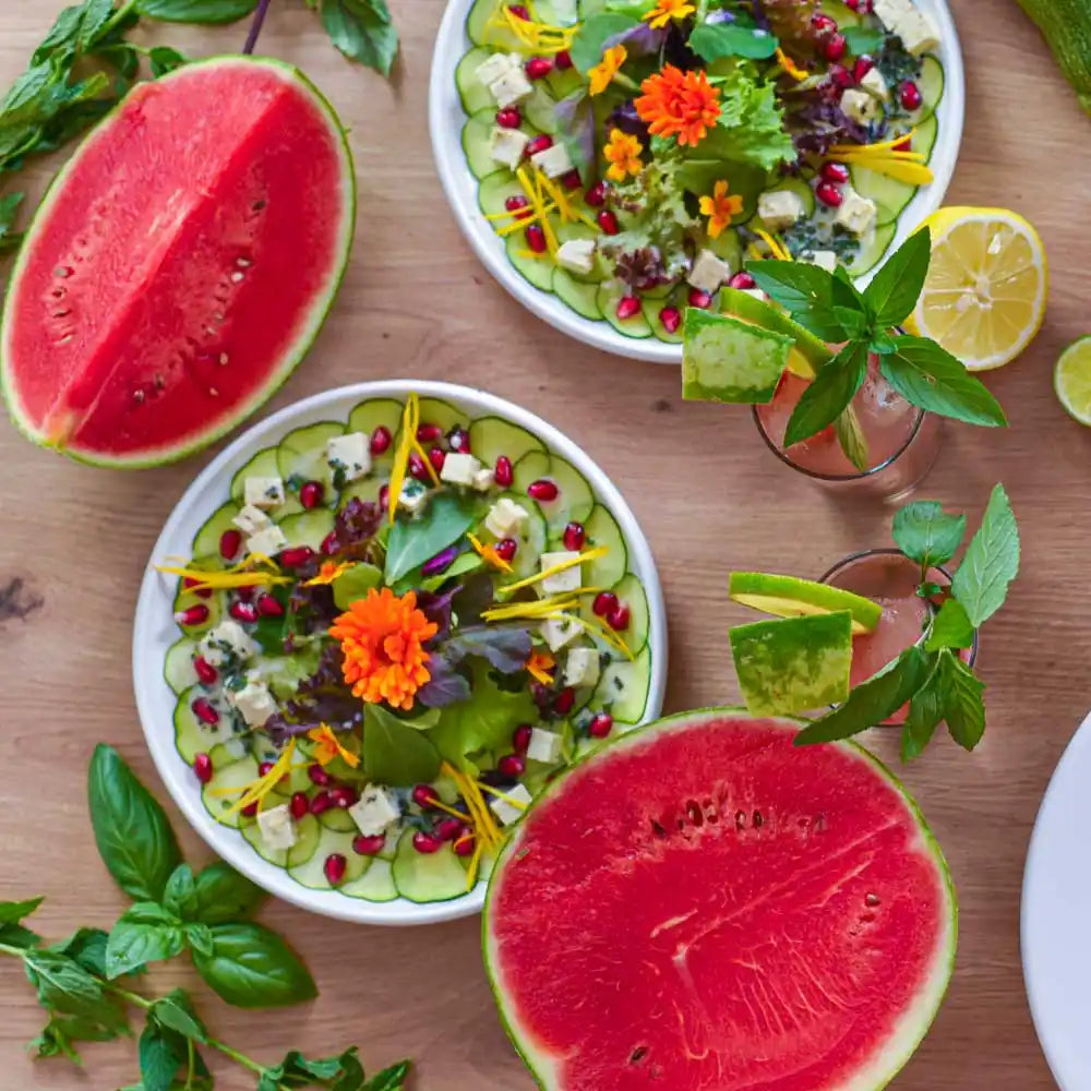 Draufsicht auf Tisch mit Gurken-Carpaccio-Salaten, halbierter Wassermelone, Drinks und frischen Kräutern.