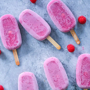 Mehrere pinke Himbeer-Eis am Stiel liegen auf grauem Untergrund, umgeben von frischen Himbeeren.