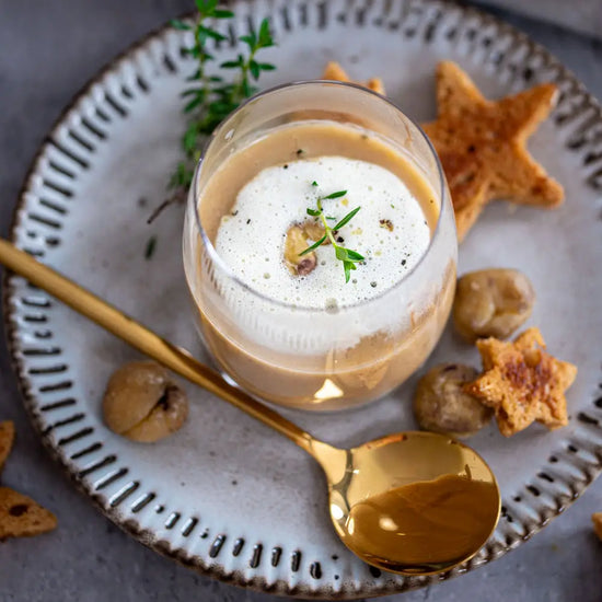 Glas mit cremiger Kastaniensuppe und Schaum, dekoriert mit Kräutern, dazu goldener Löffel und sternförmige Brotstücke auf einem Teller.