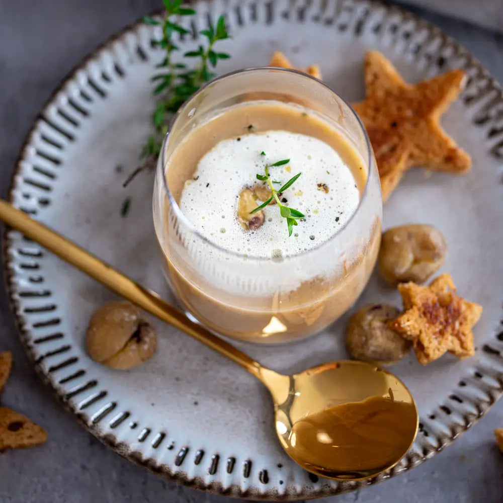 Glas mit cremiger Kastaniensuppe und Schaum, dekoriert mit Kräutern, dazu goldener Löffel und sternförmige Brotstücke auf einem Teller.