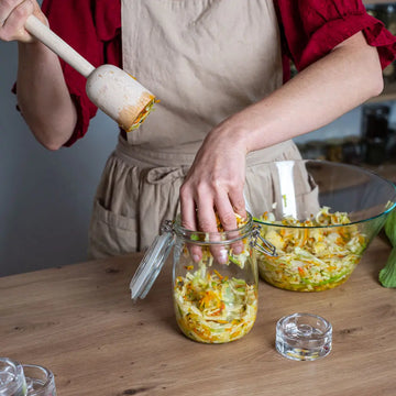 Eine Person fuellt geschnittenes Gemuese in ein 1-Liter-Fermentationsglas, waehrend sie einen Holzstampfer und ein Glasgewicht bereithaelt.